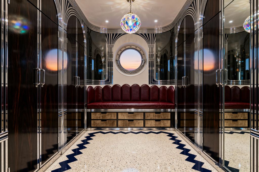 A symmetrical Art Deco mudroom lined with high-gloss dark wood wardrobes trimmed in chrome and mirrored arches. At the end, a built-in bench with a scalloped burgundy leather back sits above cane-front drawers, centered beneath a round porthole window. A terrazzo floor with a navy zigzag border leads to the seat, while an iridescent globe pendant and recessed lights reflect across the lacquered surfaces.