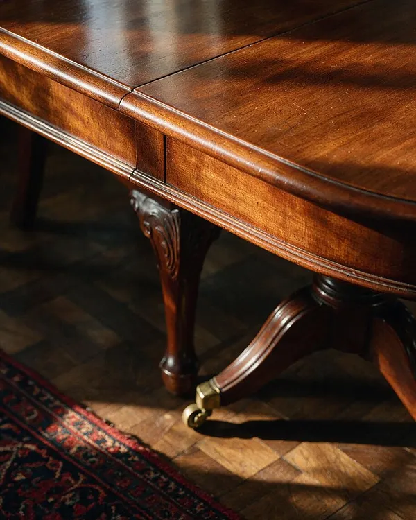 A close-up view of a traditional wooden table with a glossy mahogany finish, rounded edge molding, carved apron details, and curved legs. The table sits on a warm hardwood floor beside the edge of a red and navy patterned rug, with sunlight creating soft shadows across the wood.