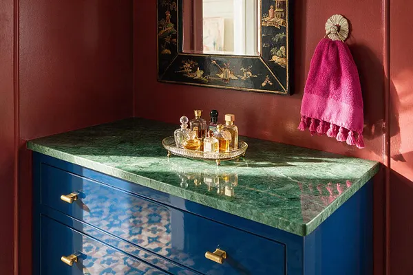 A compact vanity nook with deep red walls, a glossy blue cabinet topped by a green marble slab, and a black lacquer mirror with painted details. A bright pink hand towel hangs on the right, while a gold tray of glass perfume bottles adds sparkle on the countertop.