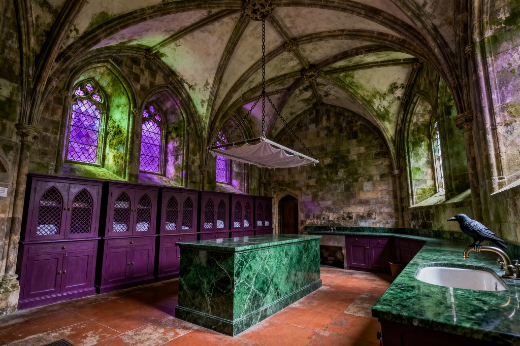A Gothic laundry room set in a ribbed-vaulted stone chamber with pointed-arch, leaded windows casting purple and green light. An emerald marble island and matching counters pair with deep plum cabinets featuring arched mesh doors holding folded linens; terracotta floor tiles, a chain-suspended drying rack, a white undermount sink with a brass faucet, and a black raven figurine complete the atmospheric, historic space.