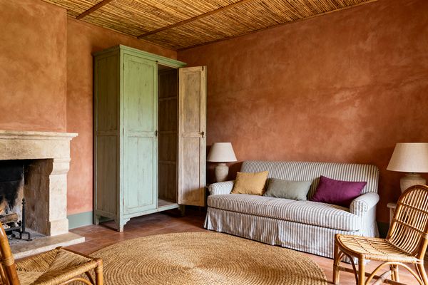 A cozy media room in French Country style features terracotta-toned plaster walls, a reed-covered ceiling, and terracotta tile floors. At left is a limestone fireplace beside a distressed sage-green armoire with one door ajar. A gray-and-white striped slipcovered sofa holds mustard, sage, and plum cushions, facing a rattan chair and a round jute rug, while two stone table lamps cast soft, ambient light.