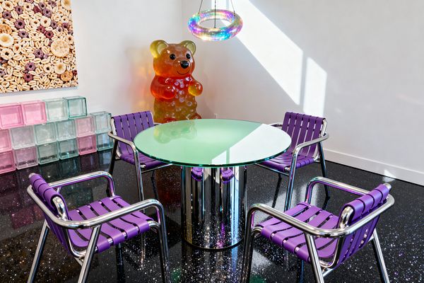 A Pop Art dining nook features a round, green-tinted glass table on a mirrored cylindrical base, surrounded by four chrome-framed chairs with purple strap seats; an iridescent ring pendant glows overhead, while a gummy bear sculpture and a large cereal photograph add playful scale. Stacked pink, mint, and clear glass blocks line the wall, with white walls and a glossy black, glitter-flecked floor illuminated by strong natural light.