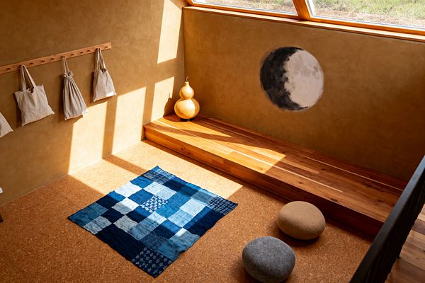 A small children’s playroom with ochre clay-plaster walls and cork flooring, lit by a sloped wood-framed window. A low built-in wooden bench runs along one wall, beside a hand-painted moon motif and a dried gourd. On the floor lies an indigo patchwork quilt with two pebble-like felt cushions nearby. A simple wooden peg rail holds neutral linen bags. The space feels minimal and tactile, using natural materials, warm light, and soft colors to create calm.