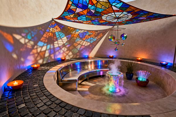 A sunken circular patio features a curved stainless-steel bench around a small fountain with a glowing, misting column that radiates rainbow light. Overhead, a geometric stained-glass style canopy casts multicolored patterns across smooth stucco walls and black cobblestone paving. Ceramic bowls with candles and subtle LED uplights add blue, magenta, and amber accents, while a planter and a bowl of fiber-optic strands complete this vivid psychedelic lounge.