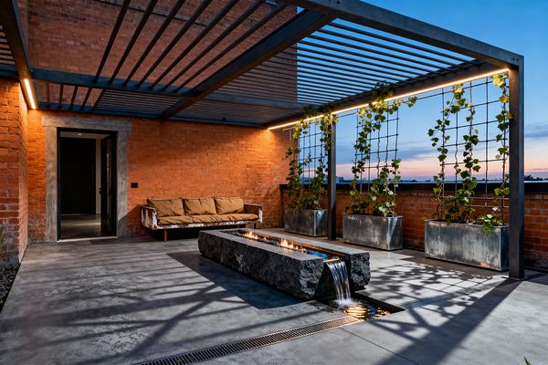 A patio with exposed red brick walls and a dark steel slatted pergola lit by linear LEDs. In the center, a long rough stone trough holds a narrow line of fire while water spills from one end into a shallow rill. The floor is smooth gray concrete with a linear drain. Along the edge, galvanized planters and wire trellises support climbing vines. A weathered metal-framed sofa with camel cushions sits near a black metal door.