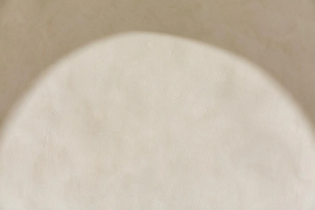 A close-up of a textured plaster or limewash wall in warm beige and cream tones, with a large curved area of sunlight creating a soft halo effect. The surface looks matte and subtly irregular, emphasizing natural texture and a calm organic modern aesthetic.