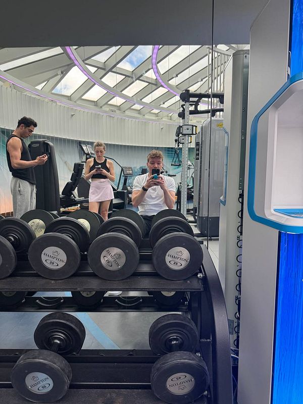 A group of three individuals is captured in a modern gym setting, with weights in the foreground and gym equipment in the background.