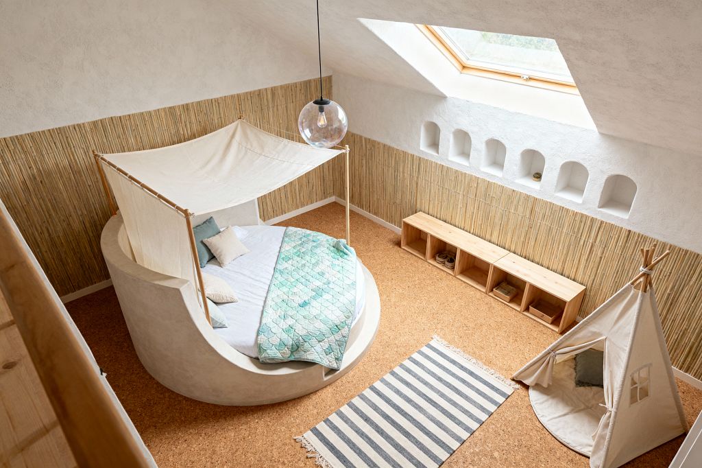 Bright kids’ room with a sloped ceiling and skylight features a circular built-in bed of pale plaster, topped by a sail-like canvas canopy and a seafoam scallop quilt. Bamboo wall cladding meets white textured plaster with small arched niches, while a pine cubby bench, gray-and-white striped rug, and a canvas teepee sit on cork flooring; a clear globe pendant hangs overhead for soft, airy light.