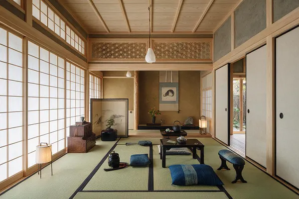 A traditional Japanese Zen-style room opens toward a backyard garden, with tatami mats, shoji screens, sliding wood-framed doors, and low tea tables. Cushions, lanterns, a hanging scroll, and natural wood finishes create a calm, minimalist atmosphere with soft daylight and a glimpse of greenery outside.