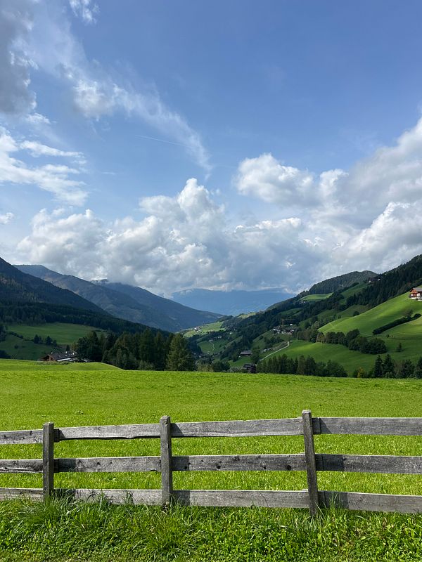 A picturesque landscape of the Dolomites featuring lush green fields and rolling hills under a partly cloudy sky.