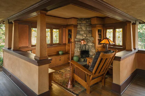 A covered Craftsman-style patio with exposed wood beams, a central stone fireplace, built-in wood cabinetry, and stained glass windows. A wooden lounge chair with a dark cushion sits on a patterned rug beside a warm amber floor lamp, creating a rustic, inviting outdoor room.