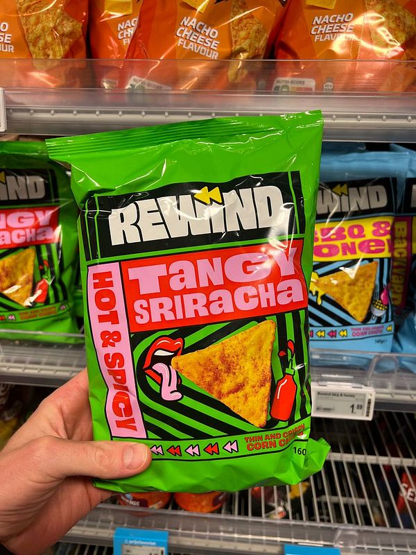 A person is holding a bag of Tangy Sriracha corn chips from the Rewind brand in a grocery store aisle.