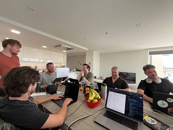 A group of six people are engaged in a collaborative work session around a table filled with laptops and snacks.
