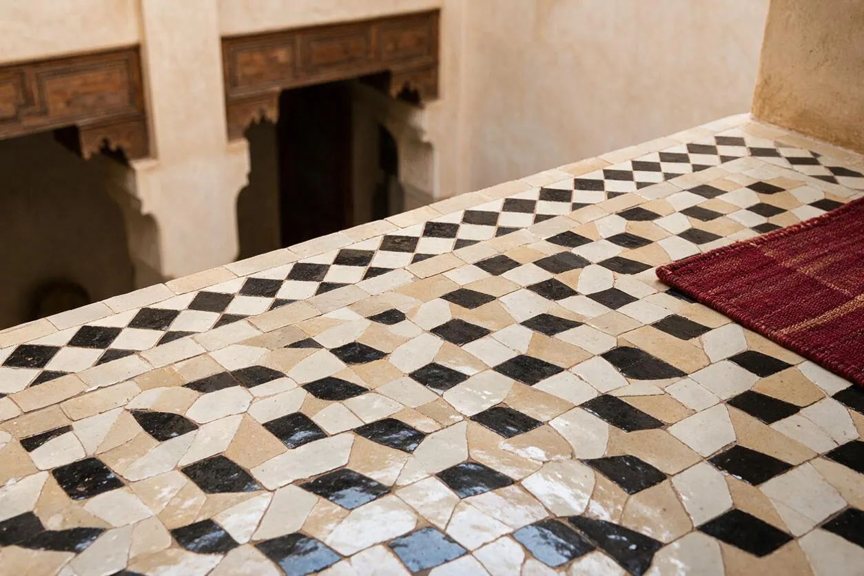 A close-up of a Moroccan mosaic tabletop or ledge made from small black, cream, and beige stone pieces arranged in a geometric pattern. In the background, a softly lit interior with carved architectural details and arched openings adds to the traditional Moroccan atmosphere.