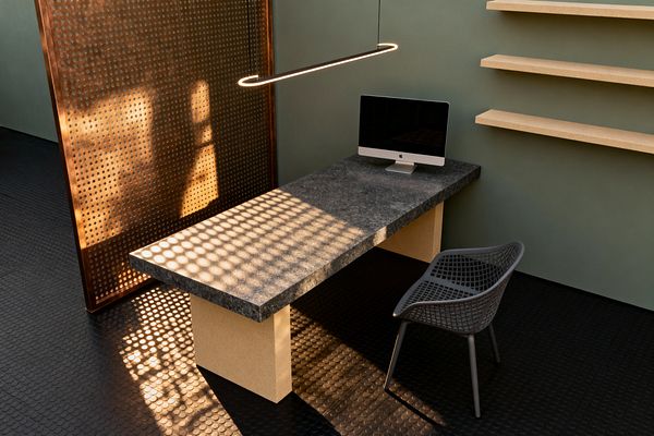 A minimalist home office with a long, thick dark stone desktop on two sand-colored block legs, holding a slim all-in-one computer. A matte black oval linear pendant hangs above, while a bronze perforated metal screen casts circular light patterns onto the desk and black rubber coin flooring. A gray mesh plastic chair sits at the desk, and three chunky beige floating shelves are mounted on a muted sage wall.