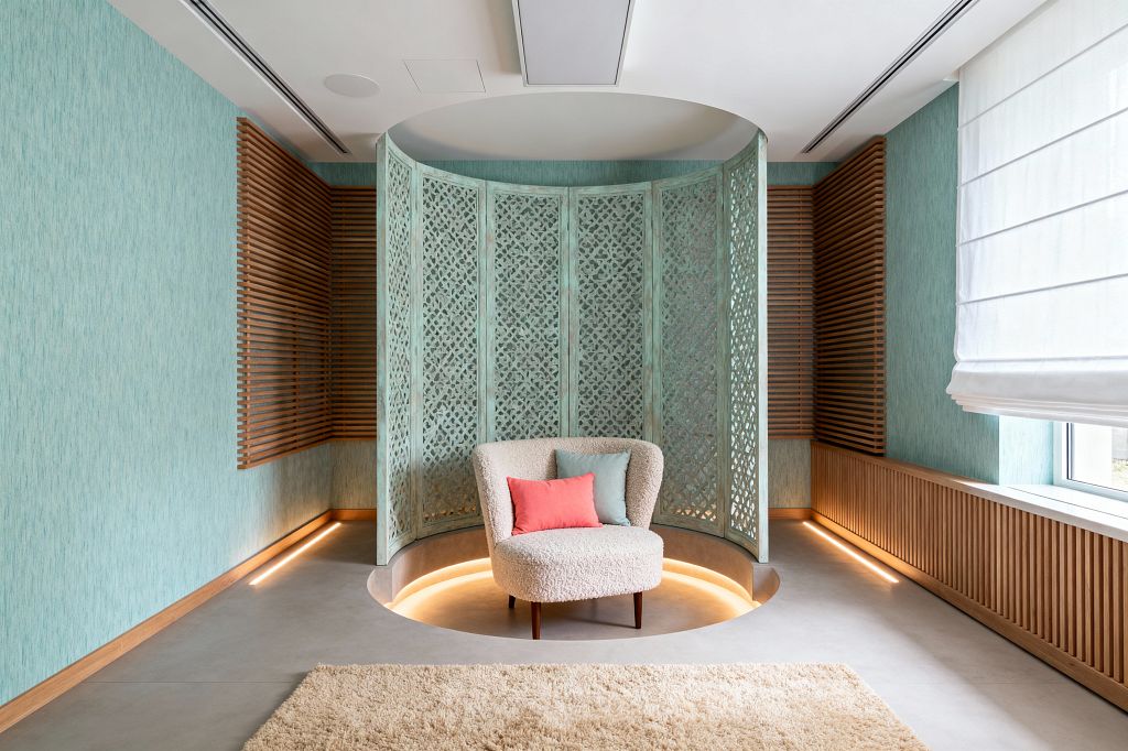 A light-filled media room with aqua walls and oak slat panels centers on a circular sunken nook enclosed by a seafoam green carved lattice screen. A cream boucle lounge chair with coral and pale teal cushions sits on a smooth microcement floor, surrounded by warm perimeter lighting, a shaggy beige rug, and a white linen Roman shade at the window, creating a calm, beach-inspired atmosphere.
