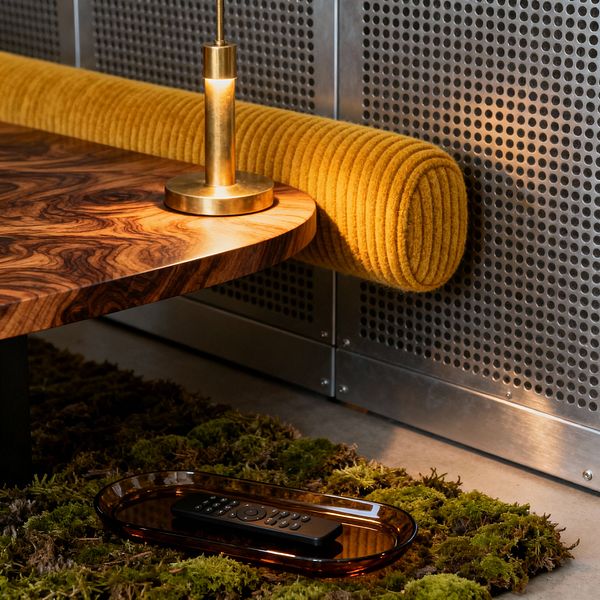 Close-up of a mid-century modern media room shows a rounded walnut coffee table with a small brass task lamp casting warm light. Behind the table, a cylindrical mustard ribbed bolster lines perforated metal wall panels, while a moss-green shag rug holds an amber glass tray with a TV remote. Warm wood, brass, and saturated yellow contrast with cool steel and concrete, creating a cozy, slightly industrial atmosphere.