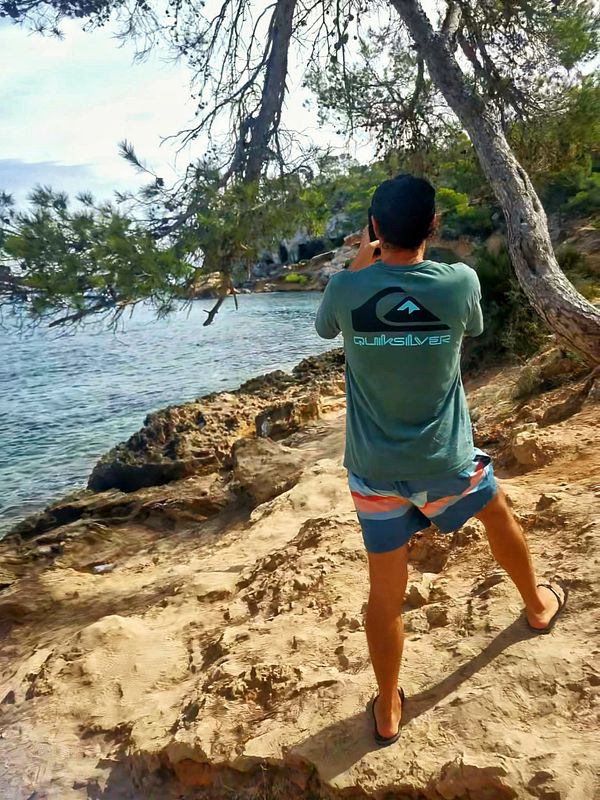A person stands on a rocky beach, taking a photo of the scenic view.