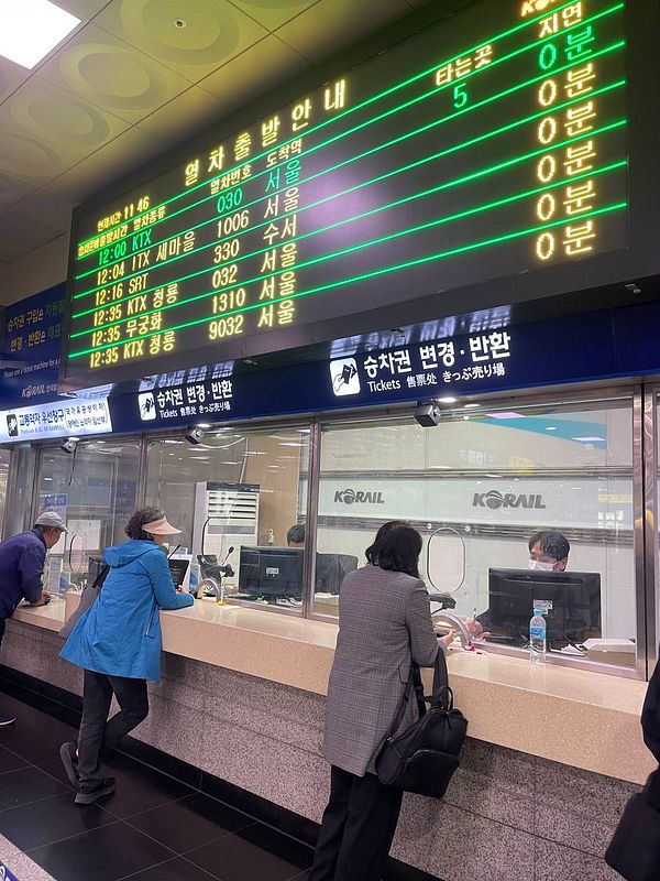A busy train station ticket counter in South Korea with a digital departure board displaying train schedules to Seoul.