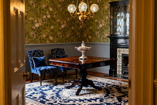 View through an open doorway into a Victorian dining room featuring a high-gloss mahogany pedestal table centered on a navy-and-cream patterned rug, with two armless side chairs in deep blue damask along the wall. The room has moss-green floral wallpaper above gray-blue wainscoting, a black cabinet and fireplace with green-and-cream tiles, a brass chandelier with three stained-glass globes overhead, and a silver tureen on the table.
