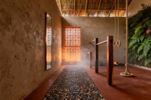 A tropical home gym with a thatched roof and textured clay-plaster walls features a pebble walkway set into a burnished red concrete floor. Sunlight filters through a terracotta lattice screen, casting warm patterns and a light mist. To the right, a living fern wall frames solid-wood parallel bars, hanging gymnastic rings, and a climbing rope. A full-length mirror reflects the space, with a kettlebell and dumbbells along the back wall.