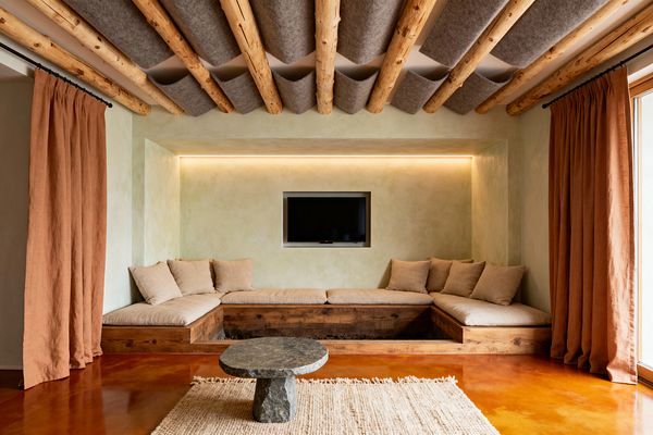 A Scandinavian-style media room features a recessed TV within a limewashed niche above a U-shaped built-in sofa with pine bases and beige linen cushions. Exposed round timber beams are spanned by gray felt acoustic panels; rust-colored linen curtains frame a large window, a rough stone round coffee table sits on a textured rug, and the warm terracotta floor glows under soft concealed LED lighting.