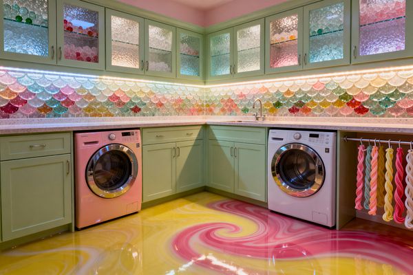 A whimsical laundry room features mint-green shaker cabinets with a light speckled countertop and glass-front uppers with textured panes. A rainbow scalloped glass-tile backsplash glows with under-cabinet lighting above a pink washer and white dryer; the glossy epoxy floor swirls yellow and pink, a small stainless sink with a chrome faucet sits in the corner, and a rod holds twisted, taffy-like textiles to complete the Candy Land theme.