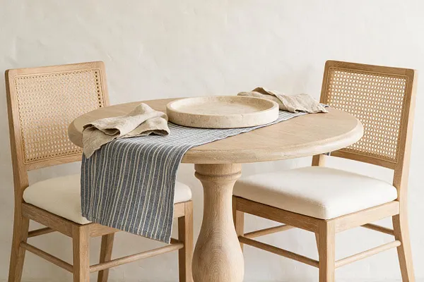 A small coastal dining vignette with a round light oak table and two matching cane-back chairs with cream cushions. A textured ceramic bowl sits on a striped blue-gray runner, with soft beige linens draped casually across the table.