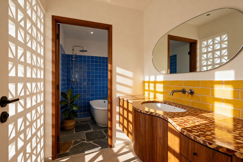 A Mid-Century Modern bathroom features a curved wood vanity with a glossy geometric countertop, a yellow brick backsplash, a large oval mirror, and a wall-mounted metal faucet. Sunlight filters through a white breeze-block screen, casting patterned shadows. Through a wood-trimmed doorway, a wet room shows cobalt blue square tiles, a freestanding white tub, a rainfall shower, slate flagstone flooring, and a potted plant in a woven basket.