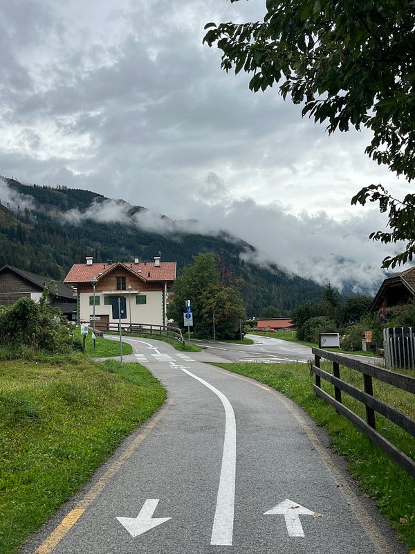 A scenic pathway leads through a quaint village surrounded by mountains and clouds.