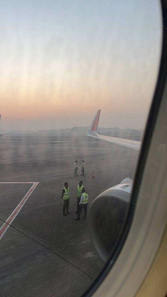 The image captures a view from an airplane window showing airport ground staff and a wing at sunrise.