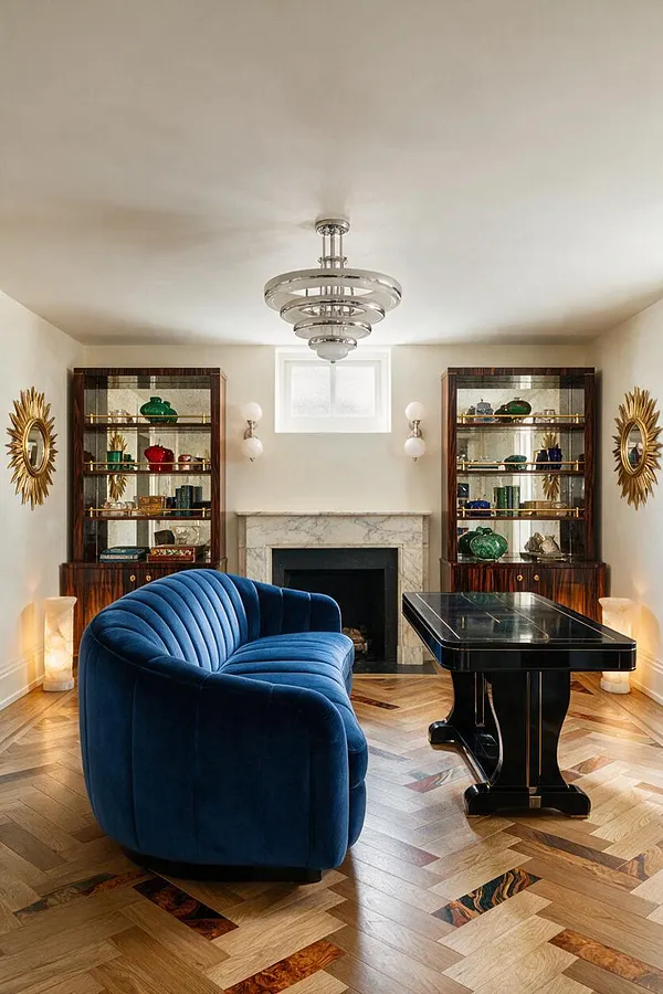 A stylish game room with a deep blue velvet curved sofa facing a glossy black game table on a herringbone wood floor. The room features a marble fireplace, mirrored wood display cabinets, gold sunburst wall mirrors, and a sculptural Art Deco ceiling light.