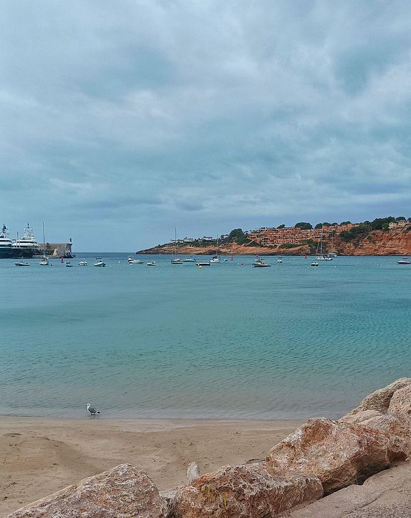 A serene coastal scene featuring a cloudy sky, calm waters, and boats anchored in the bay.