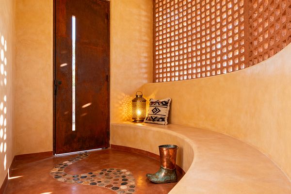A Southwestern mudroom features warm adobe-toned plaster walls and a curved built-in bench beneath a terracotta lattice screen that casts dotted sunlight. A weathered rusted-steel door with a narrow vertical window opens onto a burnished terracotta concrete floor inset with a winding river-rock mosaic; a glowing perforated metal lantern, a black-and-cream geometric pillow, and a patinated copper boot complete the cozy entry.