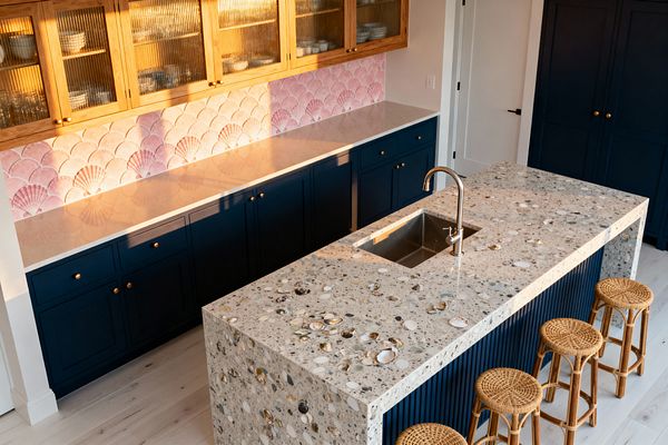 A coastal kitchen with a large island topped in shell-studded terrazzo and a fluted navy base, paired with four round rattan stools. Along the wall, deep navy shaker cabinets with brass knobs sit beneath a pale countertop and a pink scallop-tile backsplash, while upper oak cabinets with reeded glass display dishes; a stainless gooseneck faucet and undermount sink complete the sunlit, beach-inspired space.