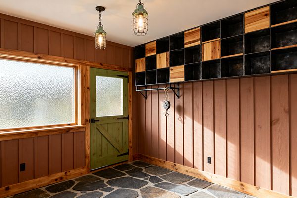 A rustic mudroom features terracotta board-and-batten walls with honey knotty-pine trim, a sage green barn-style door with a frosted glass inset, and a large privacy-glass window. The floor is irregular charcoal slate flagstone; two cage-pendant lights hang from a white ceiling, and a wall of blackened-steel cubbies with natural wood panels and a mounted rail with rope-and-pulley hook offer storage and a handmade, industrial touch.