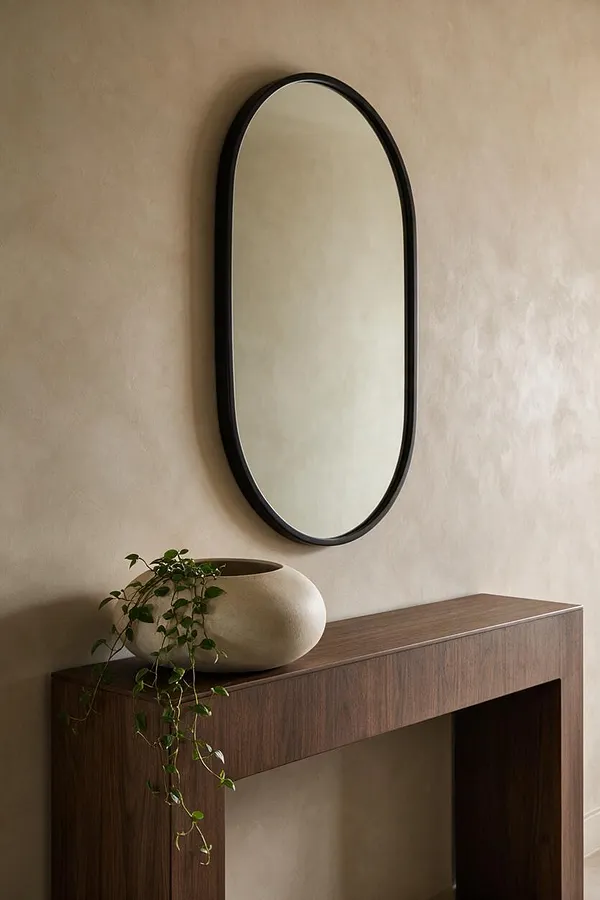 A modern entryway vignette with a tall arched mirror in a black frame mounted above a walnut console table. A sculptural cream vase with trailing greenery sits on the tabletop against a warm beige wall.