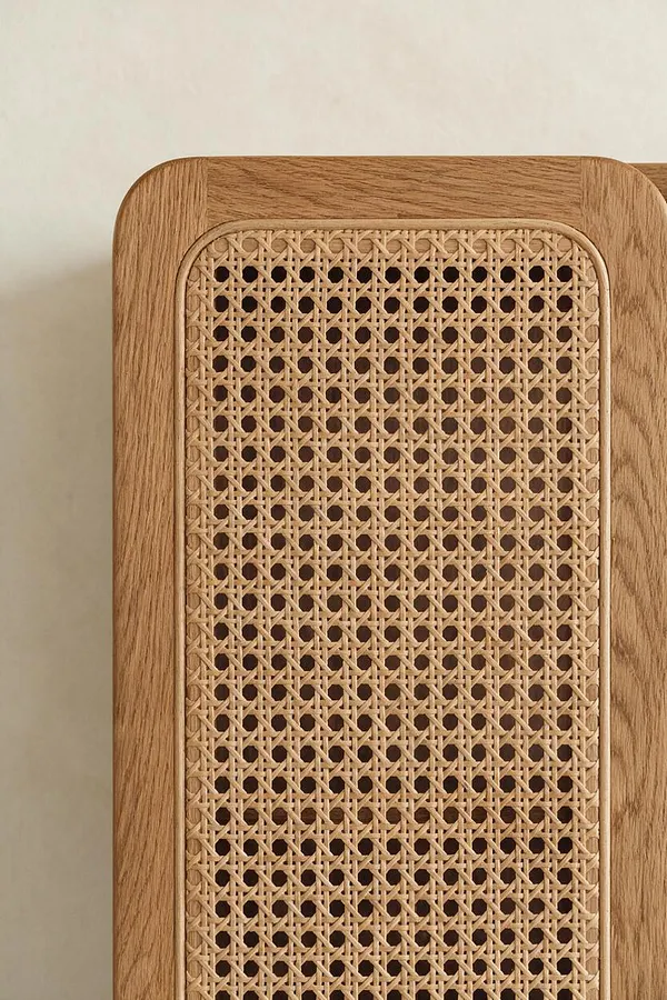 A close-up of a wooden furniture panel with rounded corners and a woven cane inset. The light oak frame surrounds a tightly patterned natural rattan lattice, set against a soft warm beige wall.