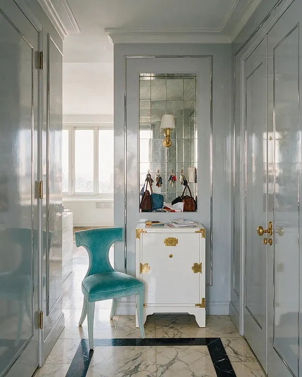 A narrow Hollywood Regency entryway with glossy pale blue paneled walls, tall mirrored trim, and brass hardware. A turquoise velvet chair sits beside a white cabinet with gold corner mounts and a wall mirror reflecting a sconce and accessories, while marble flooring with a dark border adds elegance.