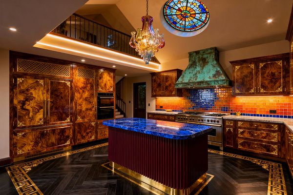 A lavish kitchen showcases burled wood cabinets with ornate brass corners and grilles, a fluted burgundy island topped by a glossy cobalt-blue stone slab, and a professional stainless range beneath a large verdigris copper hood. Orange-to-blue glazed tile, warm under-cabinet and cove lighting, a multicolored glass chandelier, a circular stained-glass skylight, and dark herringbone floors with a brass Greek-key border complete the richly layered scene beneath a mezzanine railing.