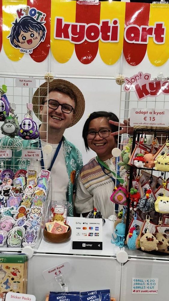 Two vendors are smiling behind their colorful art booth at an event.