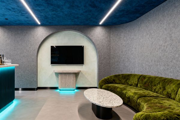 A contemporary media room features a curved moss-green velvet sectional facing a wall-mounted TV set within a pale plaster arched niche. An oval terrazzo coffee table and a fluted stone console with cyan underlighting sit on a light polished concrete floor, while gray felt acoustic wall panels and a deep cobalt-blue ceiling with linear LED strips create a softly lit atmosphere; a ribbed teal bar with a light stone top glows with matching LED at the left.