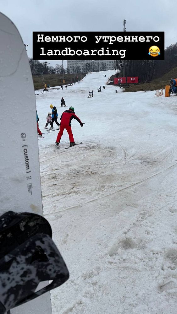 A snowy slope with people skiing and snowboarding, featuring a close-up of a snowboard in the foreground.