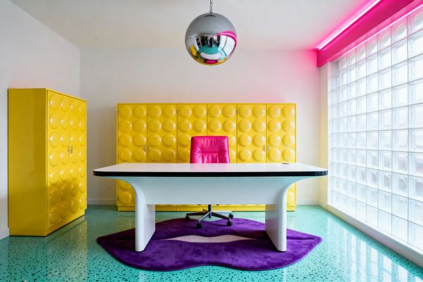 A pop art-inspired home office with a sculptural white desk over a purple organic-shaped rug, facing glossy yellow storage cabinets embossed with circular bubble panels. A hot pink desk chair, chrome globe pendant, turquoise terrazzo floor, and a wall of glass blocks lit by a magenta LED strip create a bold, playful workspace.