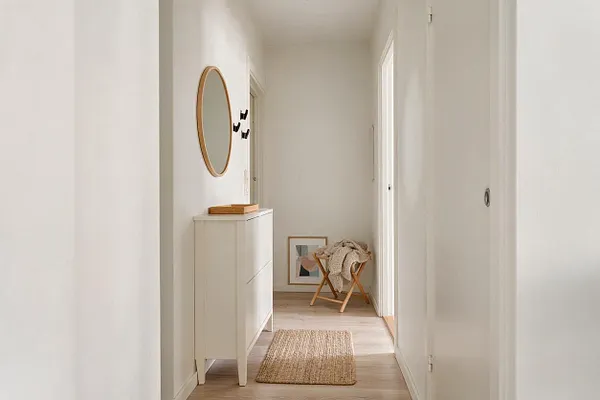 A narrow Scandinavian-style entryway with white walls, light wood flooring, and a soft neutral rug. A slim white cabinet with a round wood-framed mirror sits on the left, while a small wooden stool with a draped knit throw and a framed print add warmth at the end of the hall.