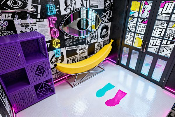 A vibrant mudroom features a bright yellow, banana-shaped bench on a polished geometric metal base, set before black-and-white comic collage walls. A vertical neon letter sign and an eye-shaped mirror hang above, with a metal rail and clear hooks beside purple perforated lockers. The glossy white coin-textured floor displays cyan and magenta footprint decals, while black French doors with Pop Art glass panels and pink LED base lighting frame the entry.