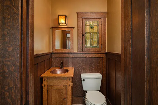 A compact Craftsman-style powder room with rich dark wood paneling and trim, a small wood vanity with a copper sink, and a white toilet. Above the vanity is a rectangular mirror and a wall sconce with warm amber light, while a built-in cabinet with a stained-glass door adds decorative detail.