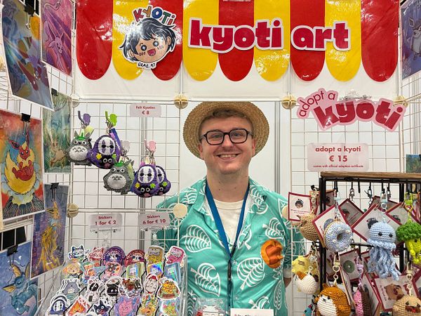 A vendor at a convention showcases their art and merchandise at a colorful booth.