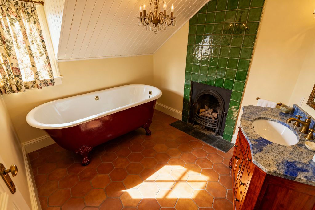 Traditional bathroom with a sloped white paneled ceiling featuring a burgundy clawfoot bathtub on terracotta hexagon tiles. A cast-iron fireplace is framed by glossy emerald green square tiles, with a slate hearth. To the right sits a solid wood vanity topped with blue-gray granite and brass fixtures. Floral curtains and a brass-and-crystal chandelier provide warm, vintage character in the cream-walled space.