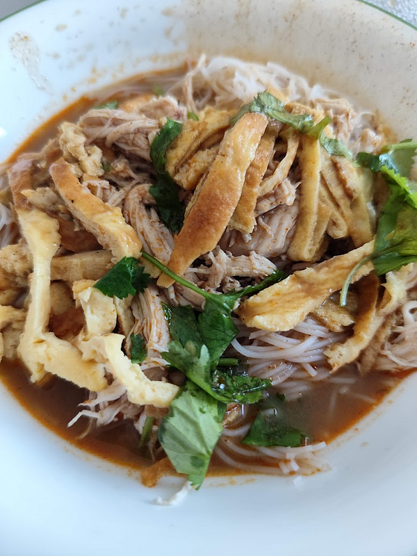 A bowl of Malaysian noodle dish featuring shredded chicken, egg strips, and fresh herbs.